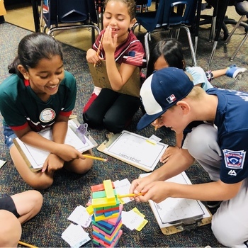 Four children sit on the floor in a classroom, smiling and laughing while playing a colorful stacking block game. Some hold clipboards and pencils, and there are scattered papers and blocks around them.