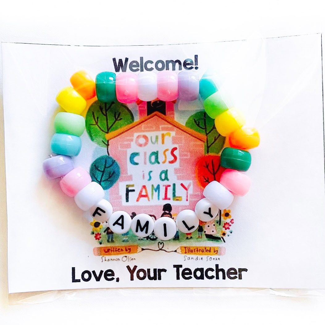 A colorful beaded bracelet with letter beads spelling FAMILY sits on a card that says Welcome! Our class is a family. Love, your teacher. The background has an illustration of a house and children.