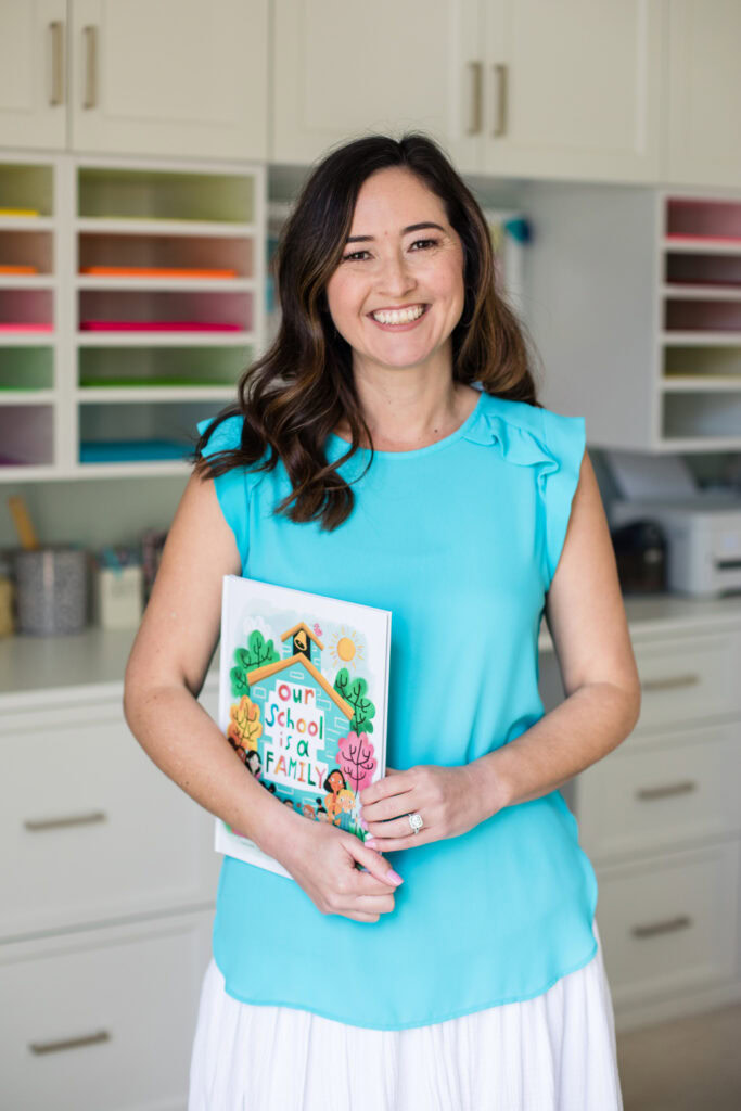 A smiling woman with long brown hair, wearing a light blue sleeveless blouse and white skirt, stands indoors holding a book titled Our School is a Family. Colorful shelves are visible in the background.