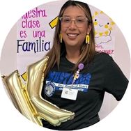 A smiling woman with glasses holding gold balloon letters K and L stands in front of classroom posters, including one that says Nuestra clase es una familia. She wears a dark shirt and a name badge.