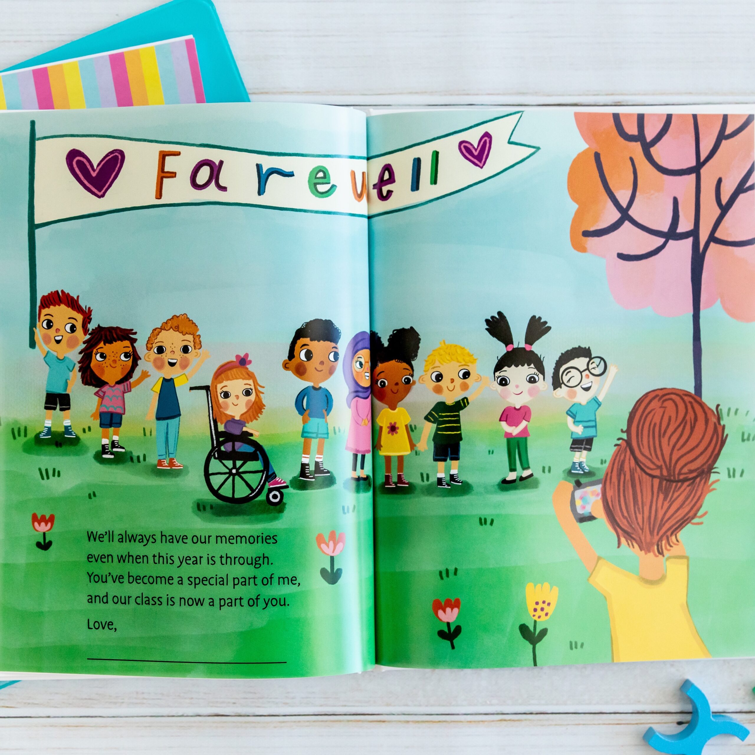 A children’s book illustration shows diverse kids standing under a Farewell banner while a girl takes their photo. The scene is set outdoors with grass, trees, and a partly cloudy sky.