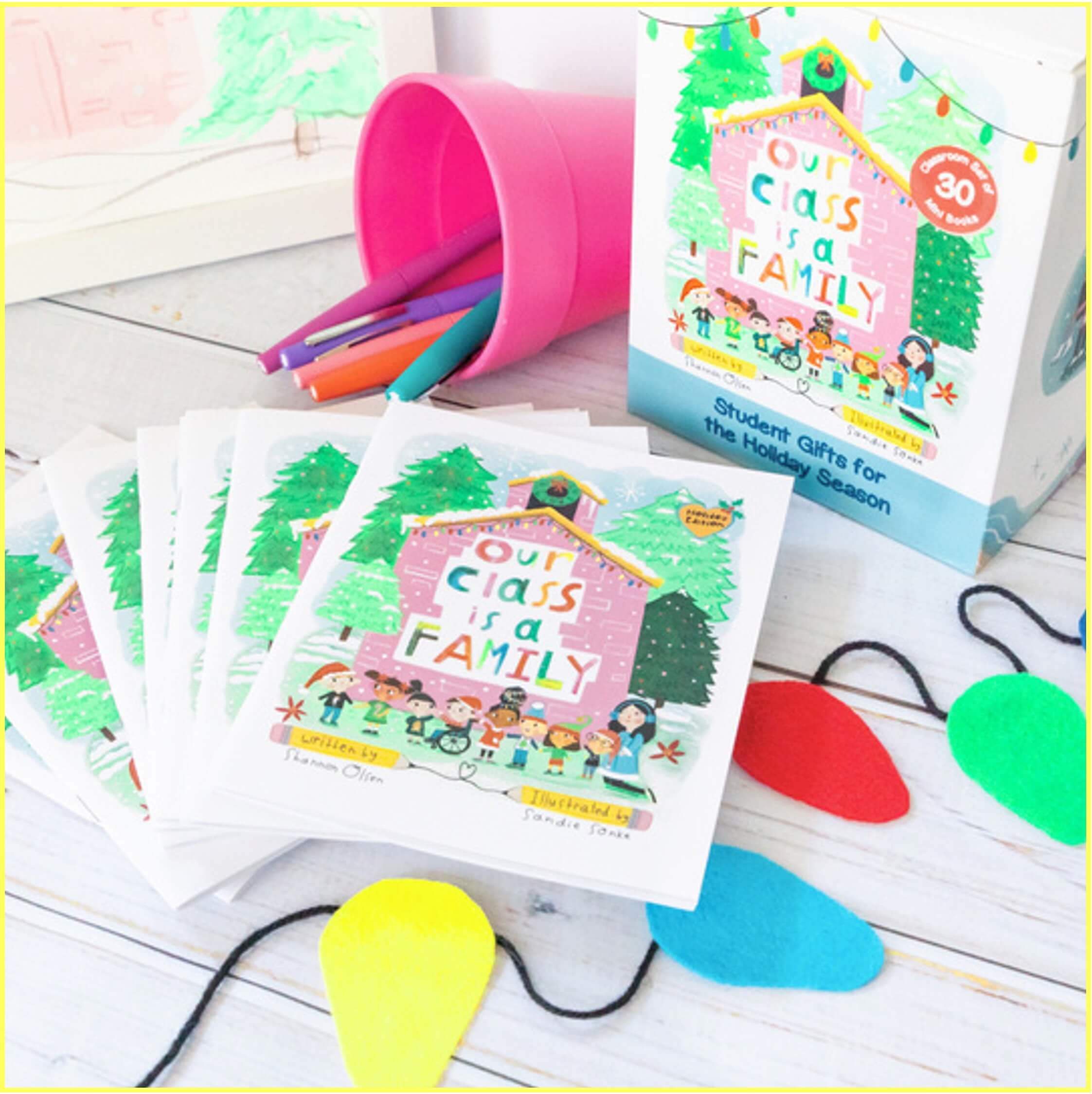 A set of illustrated books titled Our Class is a Family is displayed with colored pencils in a pink cup, colorful felt cutouts, and a decorated box labeled as a holiday student gift.
