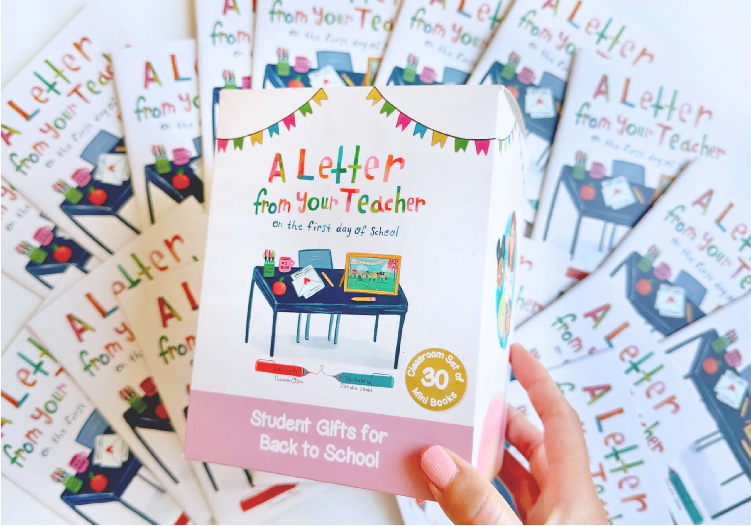 A hand holds a box labeled A Letter from Your Teacher on the First Day of School with colorful illustrations. Multiple copies of the same book are spread out underneath the box.
