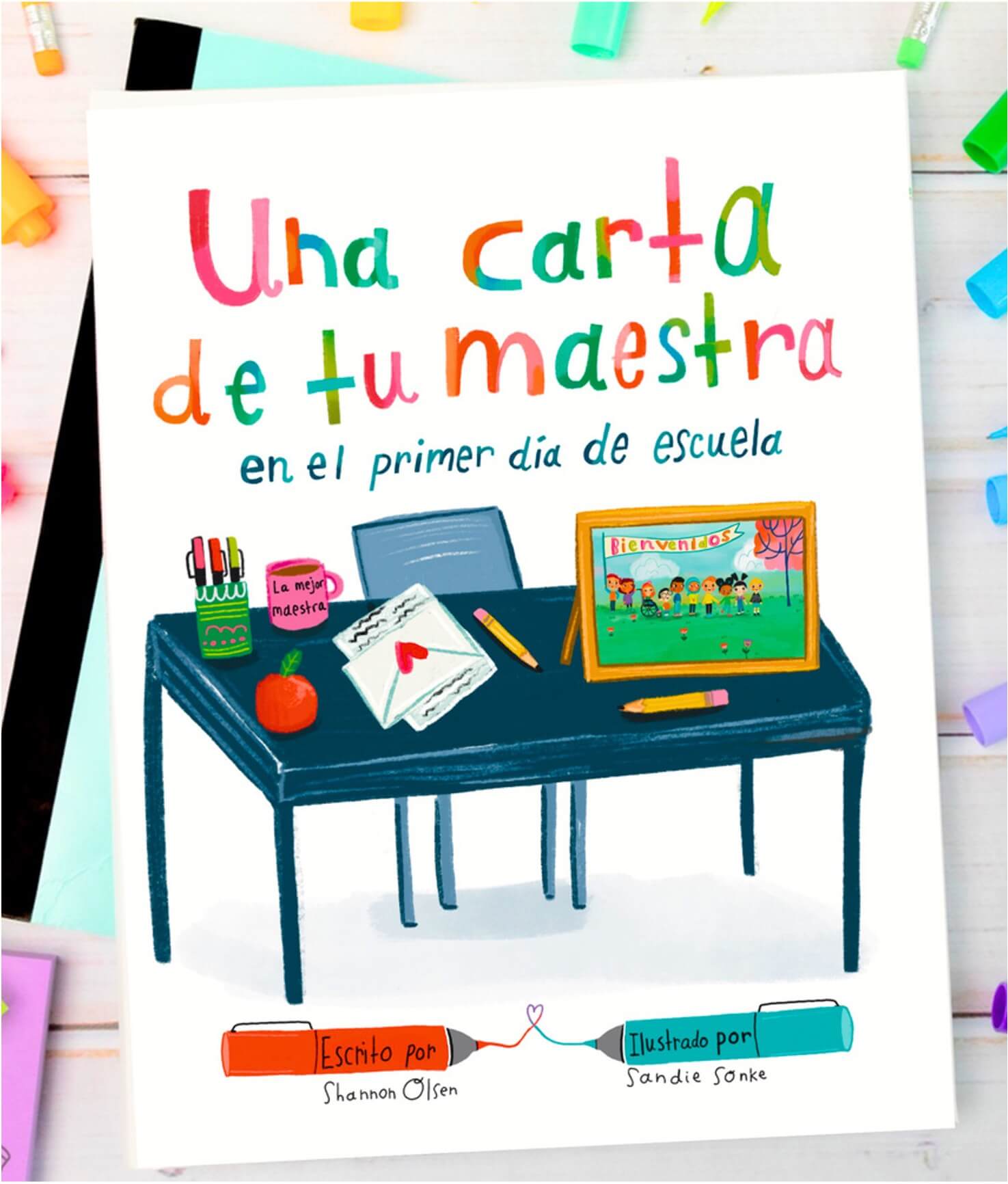 Book cover titled Una carta de tu maestra en el primer día de escuela shows a classroom scene with a desk holding an envelope, apple, mug, and a chalkboard with a welcome drawing. Bright, colorful text and accents.