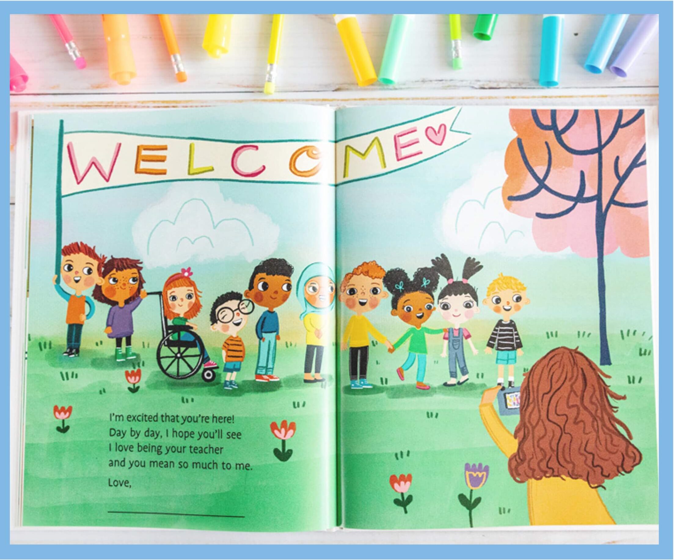 An illustrated childrens book shows a teacher taking a photo of a diverse group of smiling children standing under a WELCOME banner in a park. Colorful markers are arranged above the open book.