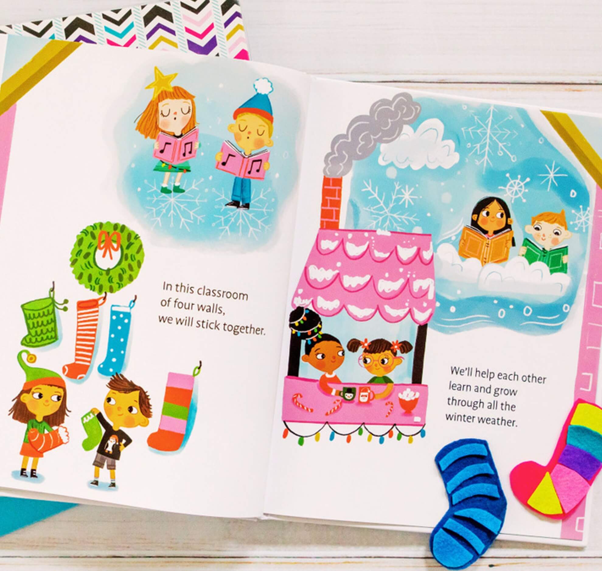 Colorful illustration of children singing, hanging stockings, and reading inside a classroom. Two children look out a frosty window as snow falls. Holiday decorations and warm winter clothing fill the scene.