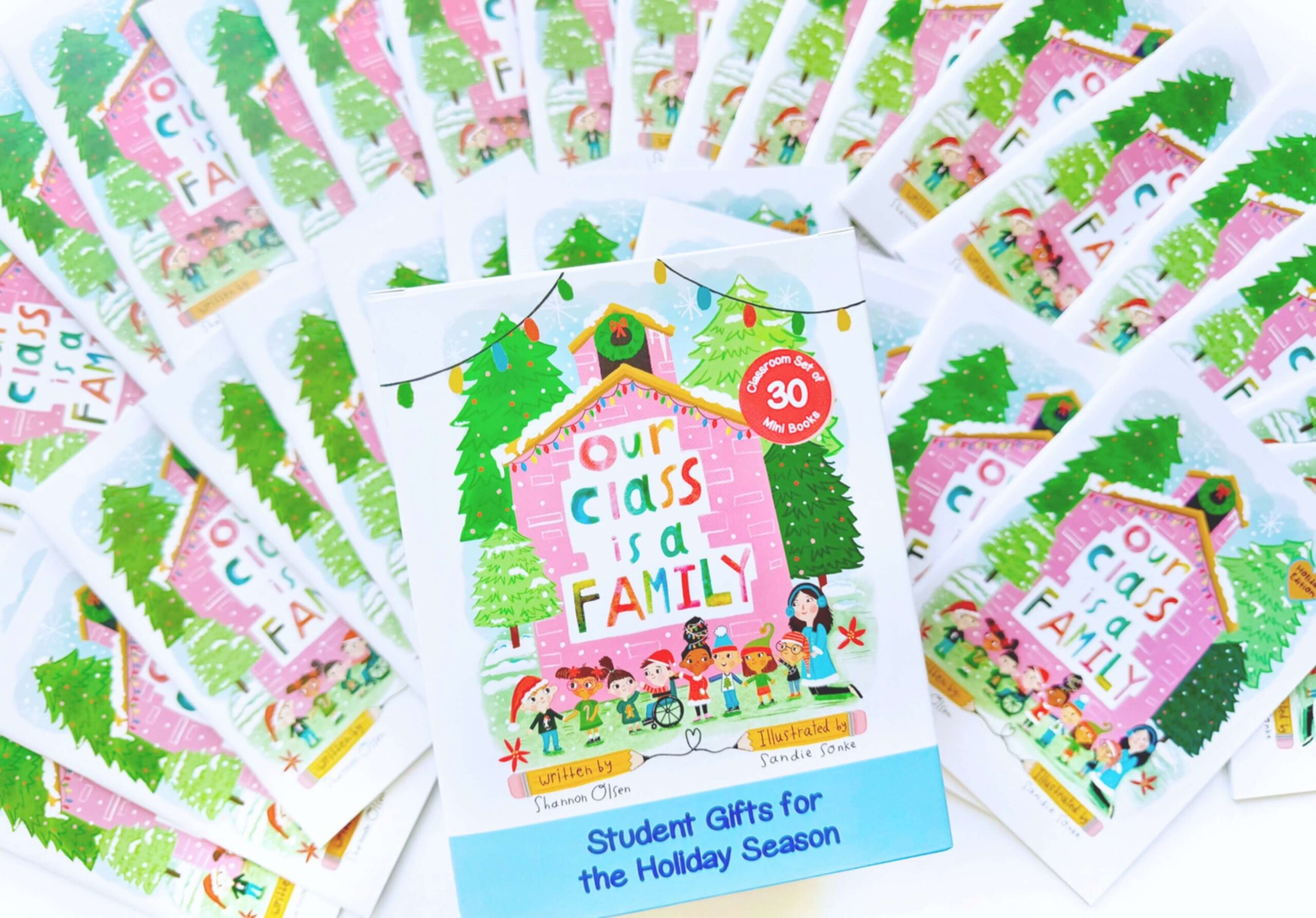 A display of Our Class is a Family books arranged in a fan shape, with one book in the center featuring colorful illustrations of children, a pink house, and holiday decorations.